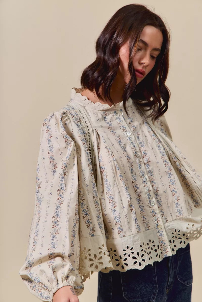 Woman wearing a cream button-down blouse with delicate floral print, lace trim, and eyelet cutout hem, styled with dark denim jeans and a tan hat.