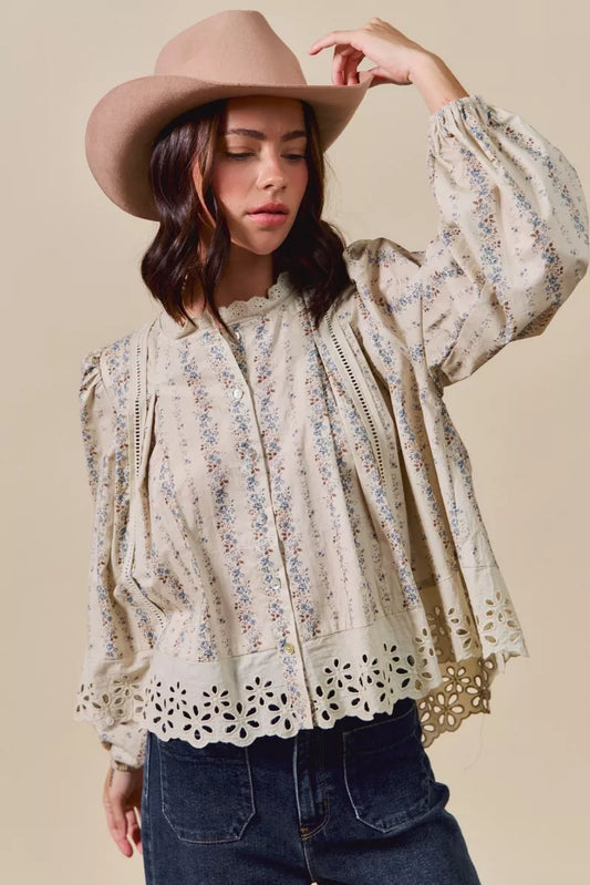 Woman wearing a cream button-down blouse with delicate floral print, lace trim, and eyelet cutout hem, styled with dark denim jeans and a tan hat.
