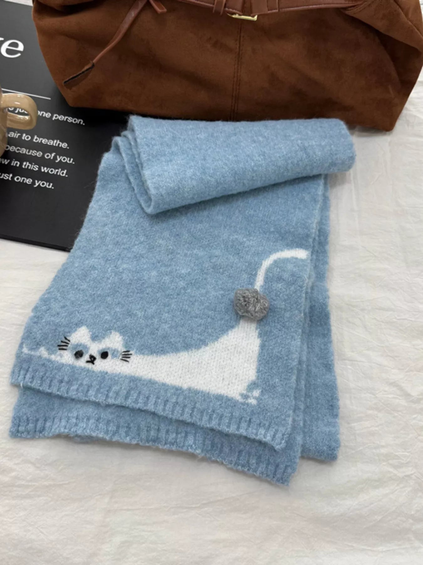 Soft knit scarf with a playful cat design and pom-pom detail styled flat on fabric background