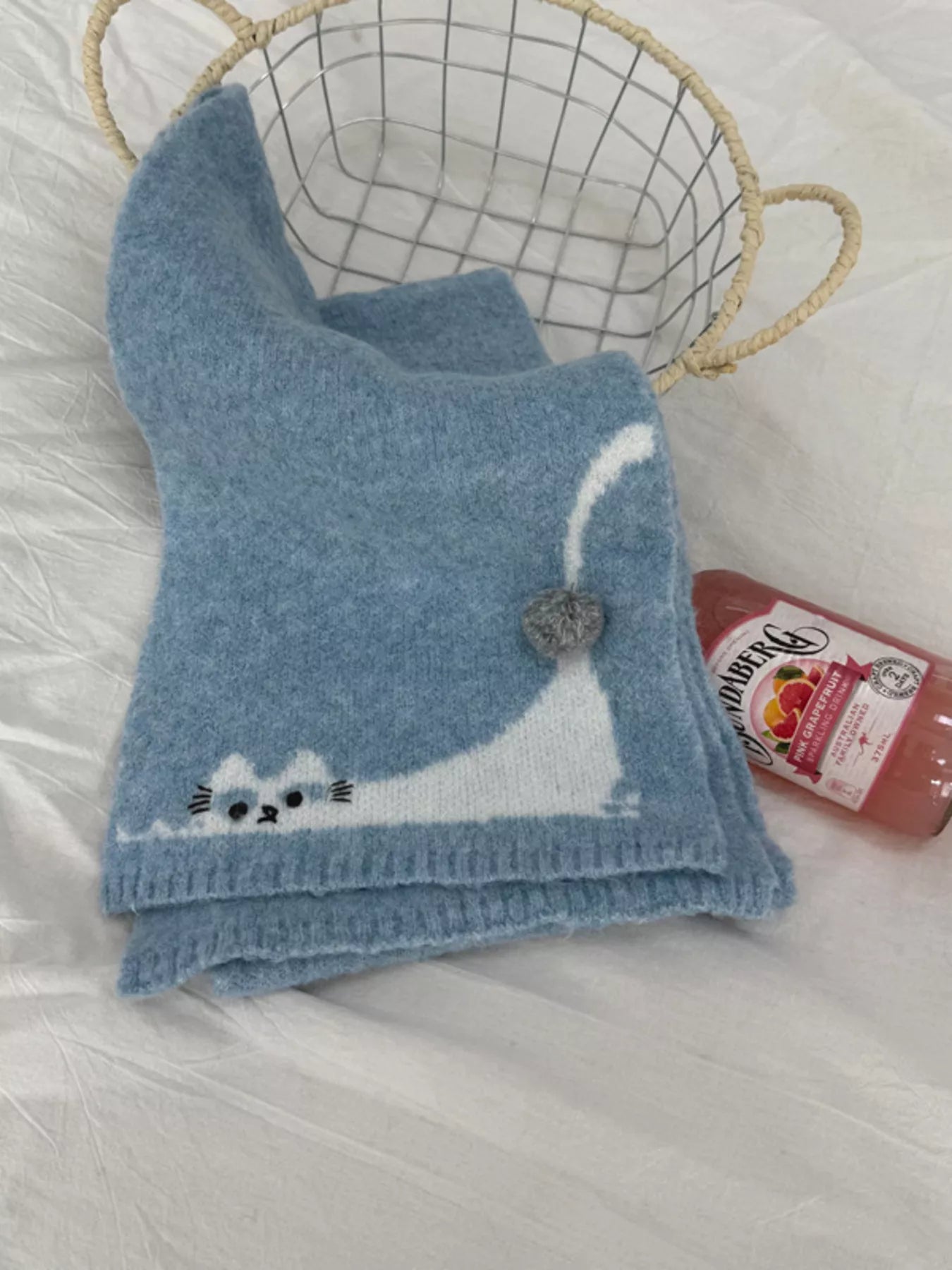 Soft knit scarf with a playful cat design and pom-pom detail styled flat on fabric background