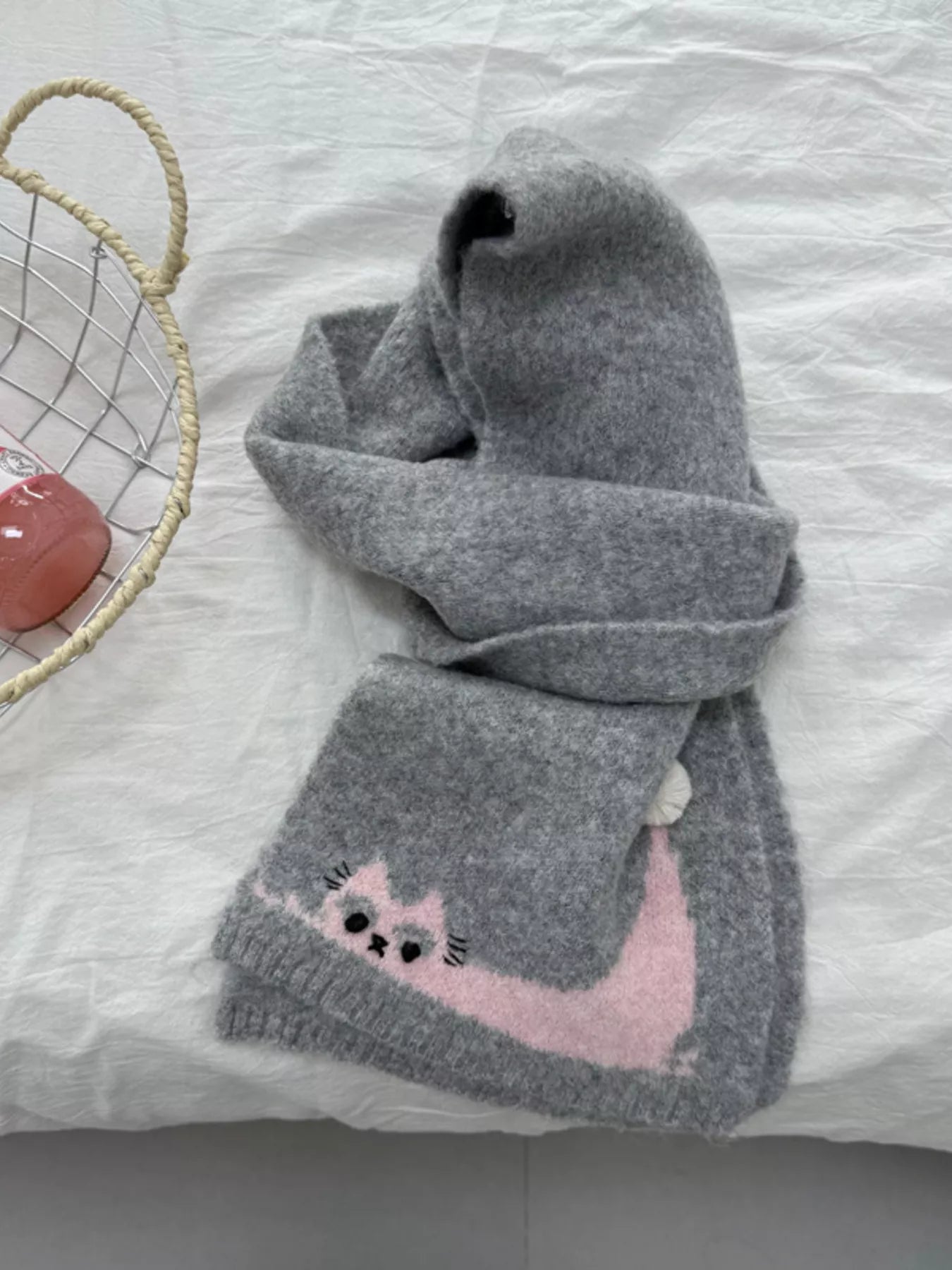Soft knit scarf with a playful cat design and pom-pom detail styled flat on fabric background