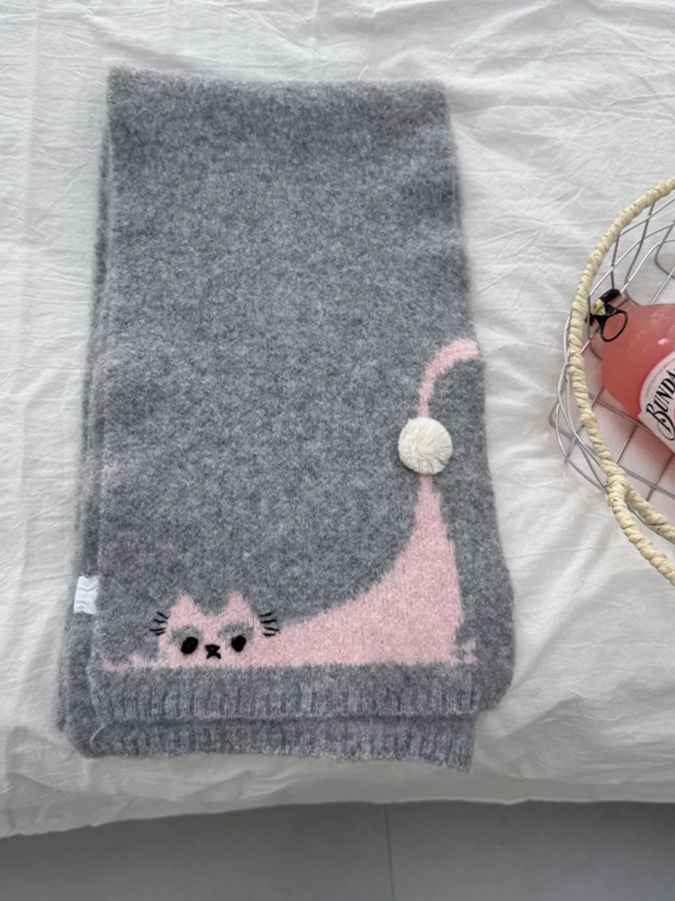 Soft knit scarf with a playful cat design and pom-pom detail styled flat on fabric background