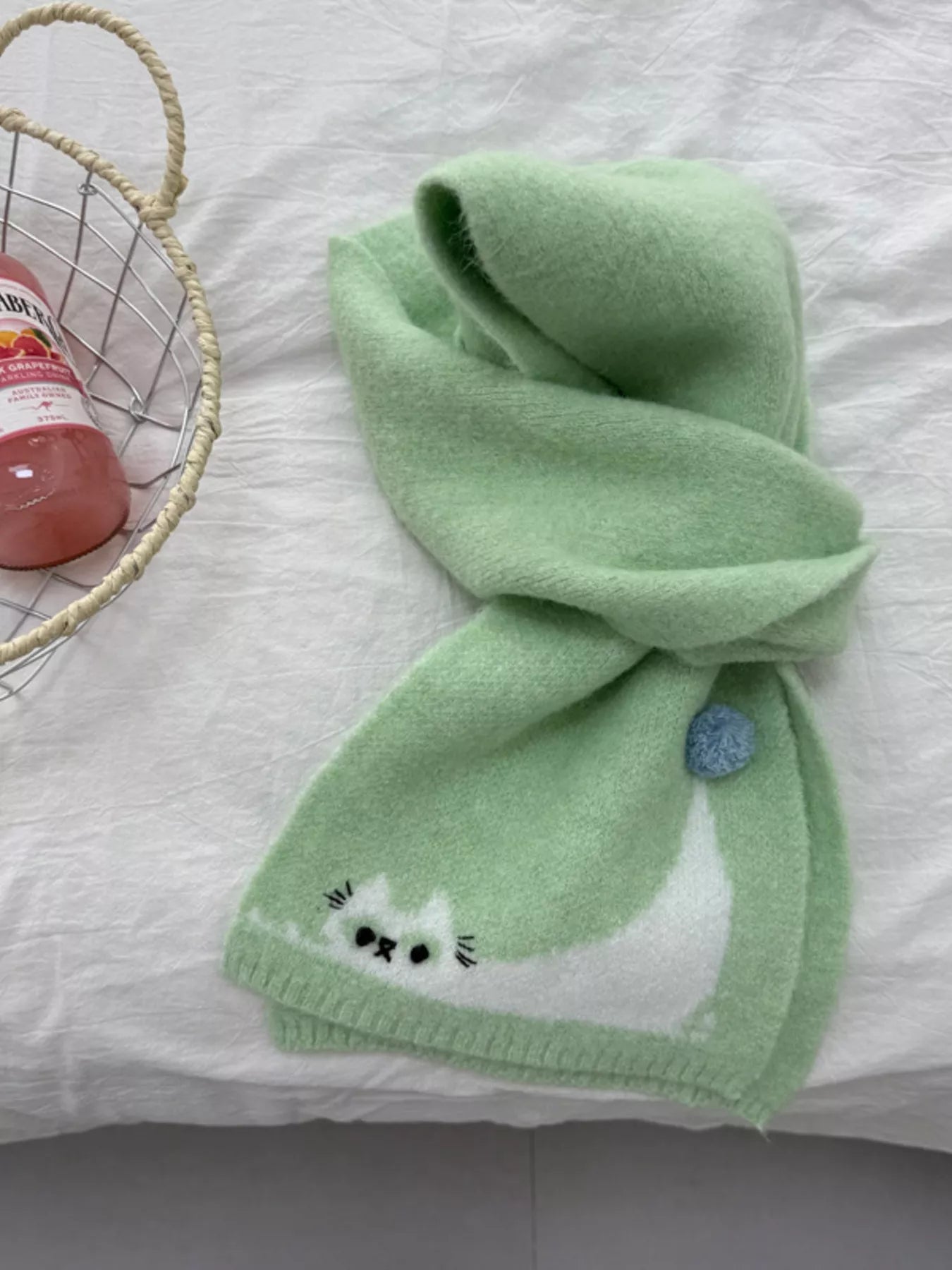 Soft knit scarf with a playful cat design and pom-pom detail styled flat on fabric background