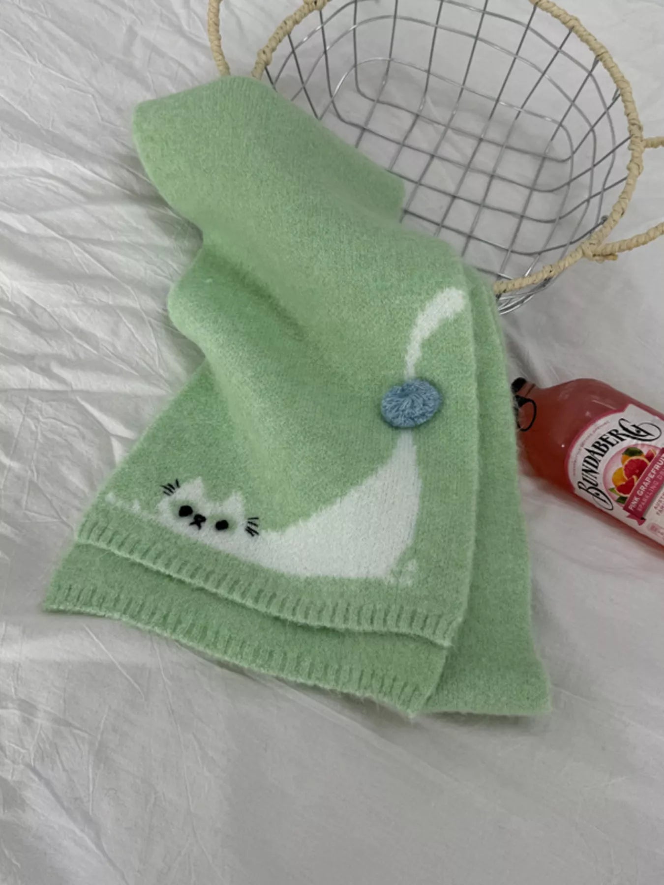 Soft knit scarf with a playful cat design and pom-pom detail styled flat on fabric background