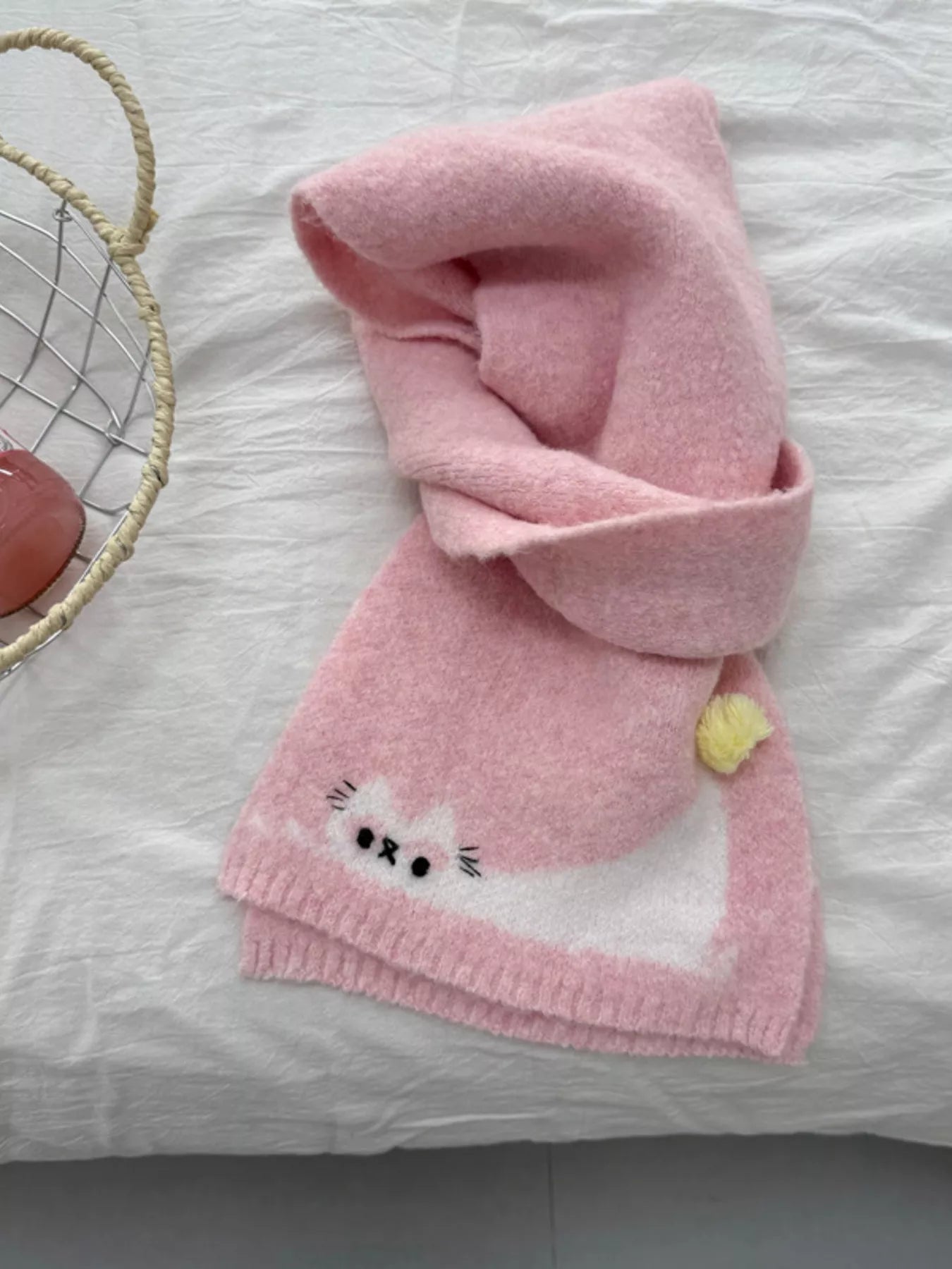 Soft knit scarf with a playful cat design and pom-pom detail styled flat on fabric background