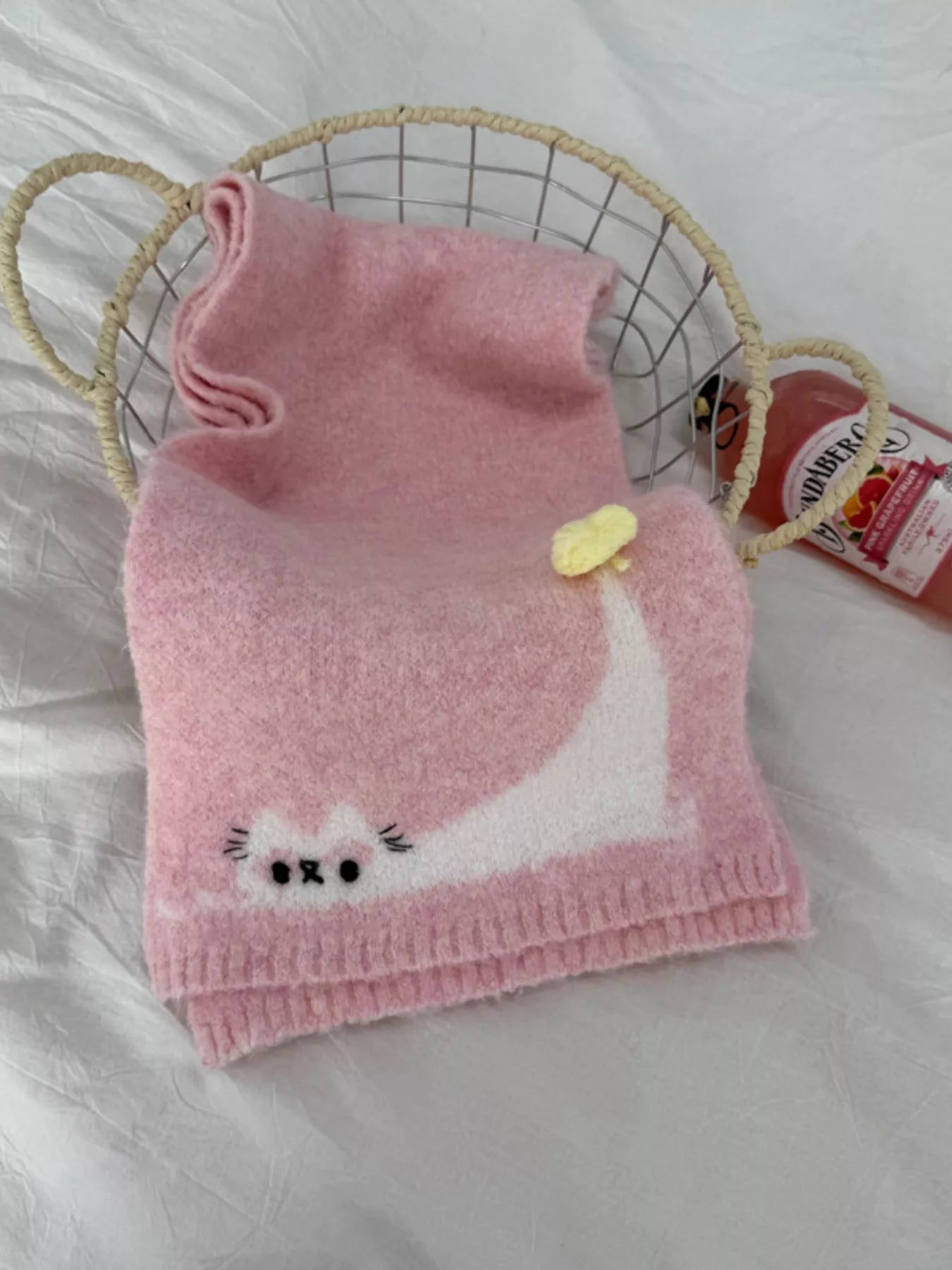 Soft knit scarf with a playful cat design and pom-pom detail styled flat on fabric background