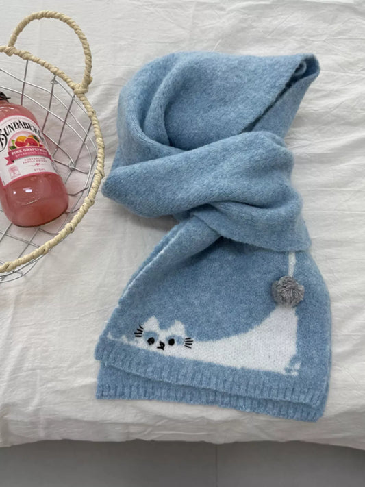Soft knit scarf with a playful cat design and pom-pom detail styled flat on fabric background
