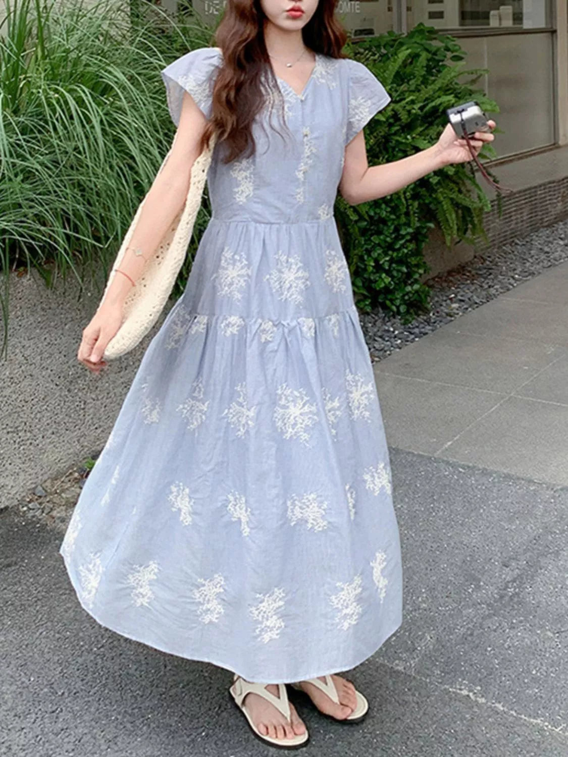 Model wearing the Whispering Bloom Buttoned Maxi Dress in light blue with white floral print, short sleeves, and a flowy tiered design.