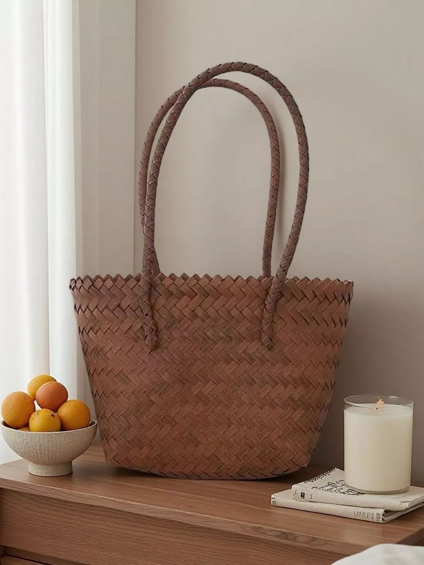 Medium woven brown tote bag with braided handles, displayed on a wooden surface beside a bowl of oranges and a lit candle.