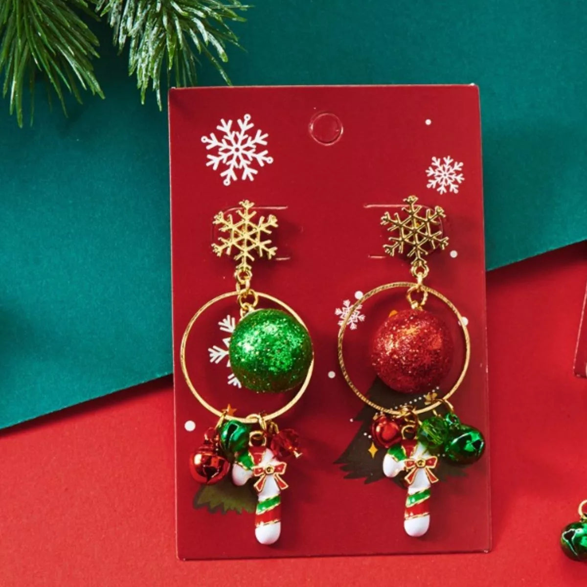 Gold snowflake holiday earrings with green and red glitter baubles, tiny bells, and candy-cane charms displayed on a red festive card.